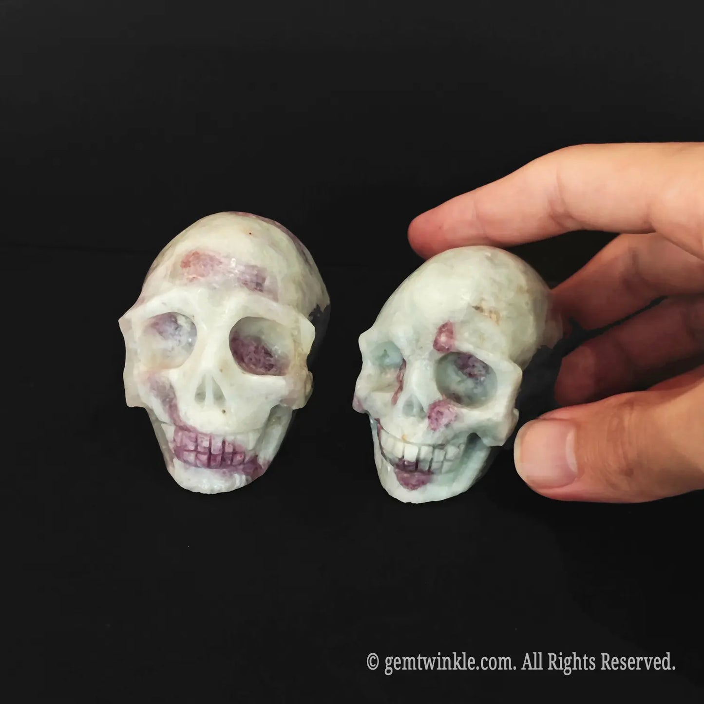A hand adjusting two 6cm pink tourmaline in quartz human skulls to inspect their anatomical features