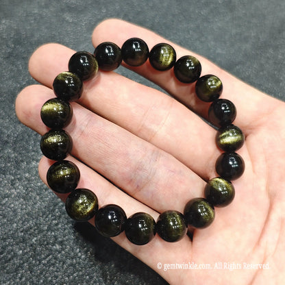Fingertips holding a natural gold sheen obsidian stretch bracelet showcasing smooth polish and optical depth