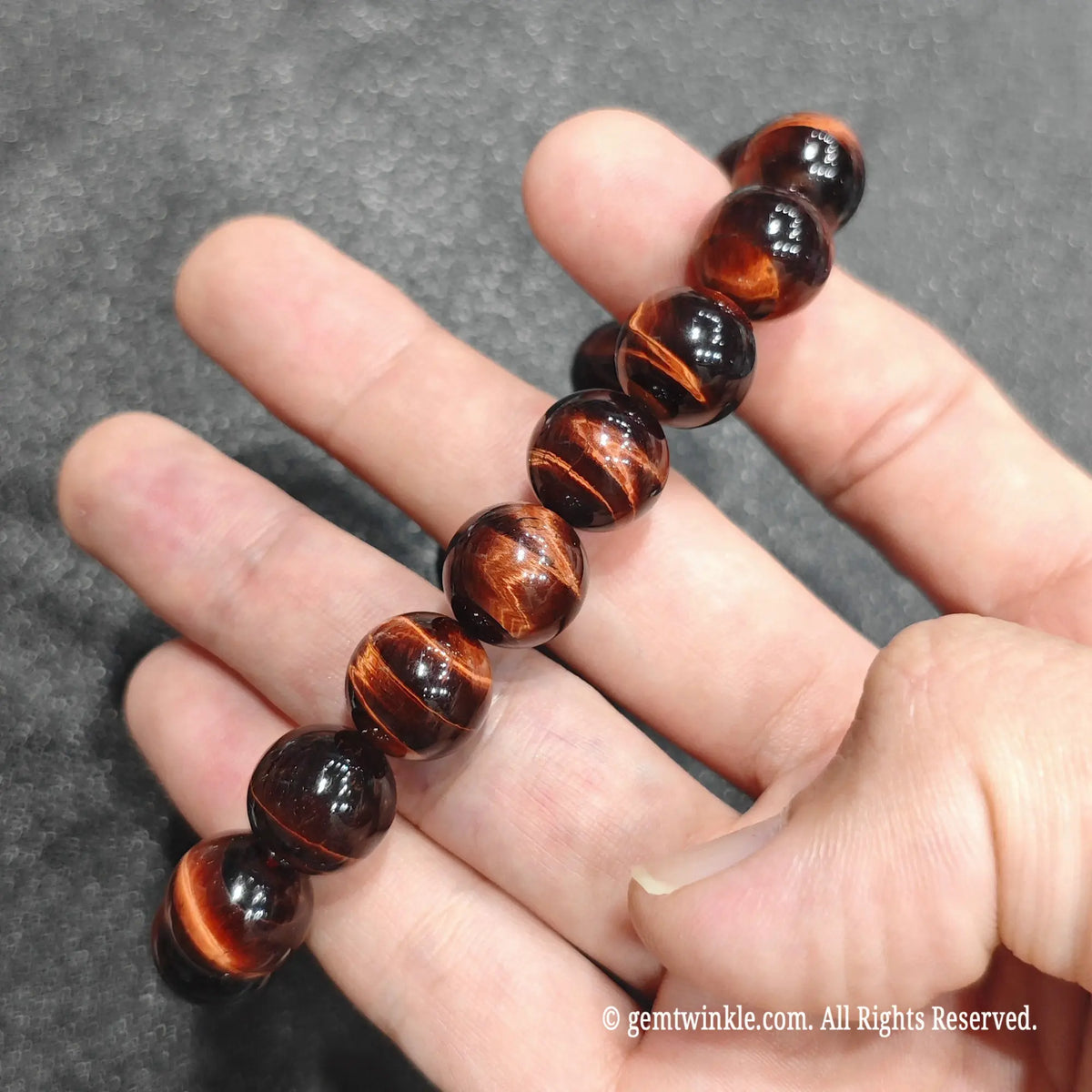 Fingertips holding a 12mm red tiger's eye beaded wristlet displaying sharp parallel lines of light reflection
