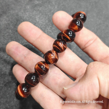 Fingertips holding a 12mm red tiger's eye beaded wristlet displaying sharp parallel lines of light reflection