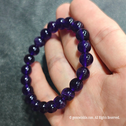 Fingertips holding an 8mm deep amethyst beaded bracelet displaying polished luster under ambient studio light[cite: 8]