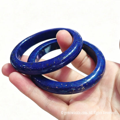 Close up view of natural lapis lazuli solid bangles held between fingers displaying high gloss finish and golden flecks