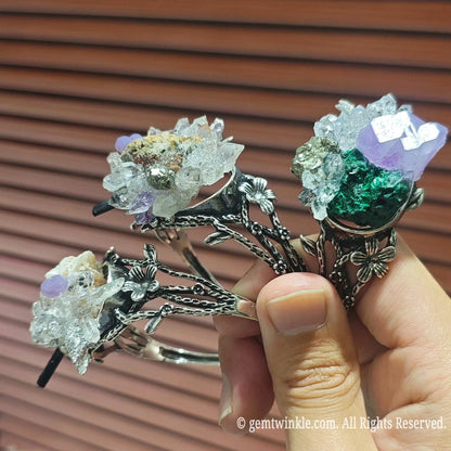 Hand holding two raw crystal cluster adjustable cuff bracelets displaying random mineral combinations and quartz points
