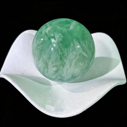 Backlit green fluorite sphere revealing ethereal white feather inclusions and soft glow through translucent stone