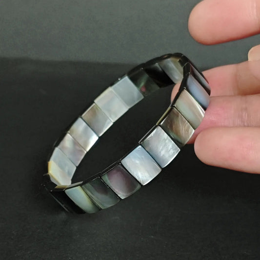 Natural black mother-of-pearl rectangular bead bracelet held in hand, showing green-blue iridescence and smooth faceted edges on dark slate background