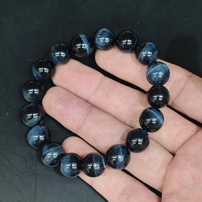 Hand holding a classic blue tiger’s eye bracelet with consistently sized round beads (approx. 8–10mm). Deep indigo-black base with vivid electric-blue bands that reflect light sharply. Elastic fit, no metal.