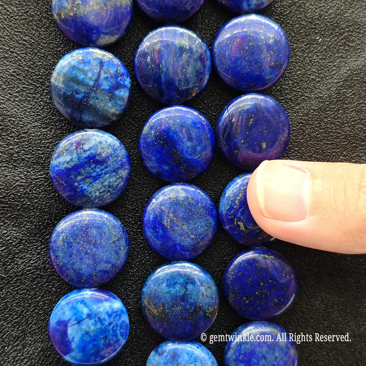 Finger pointing to a smooth flat round natural lapis lazuli coin bead displaying white calcite veins and metallic golden pyrite sparkles