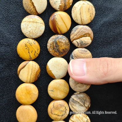 Finger pointing to a smooth flat round picture jasper coin bead displaying earthy brown and cream landscape lines