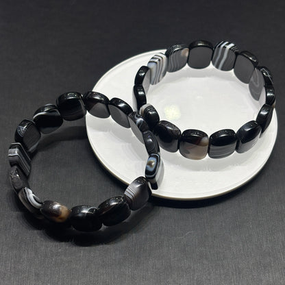Black and white banded onyx square bead bracelets crossed on dark surface, stylish aesthetic casual wear jewelry