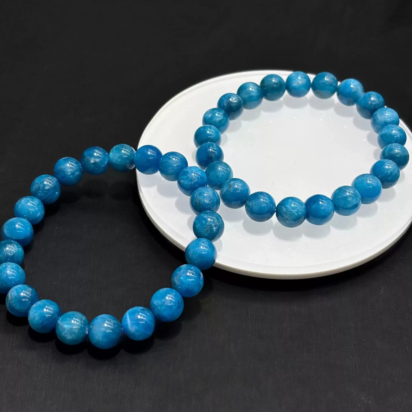 Two natural blue apatite bracelets overlapping on white dish under soft studio light, emphasizing depth and clarity of the stone