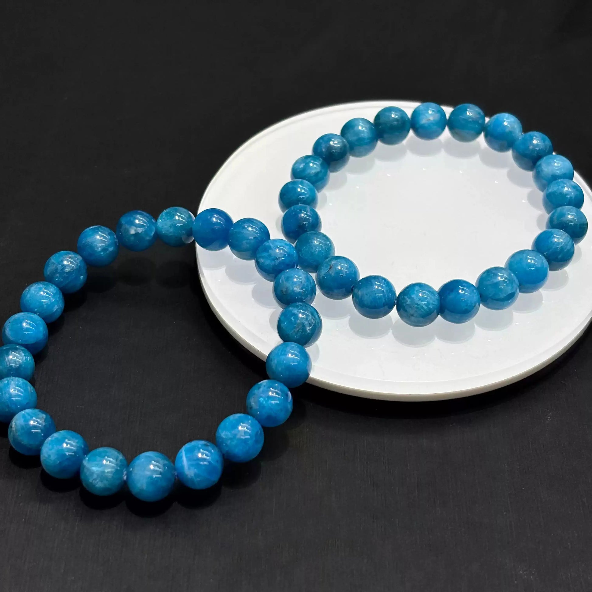 Two natural blue apatite bracelets overlapping on white dish under soft studio light, emphasizing depth and clarity of the stone