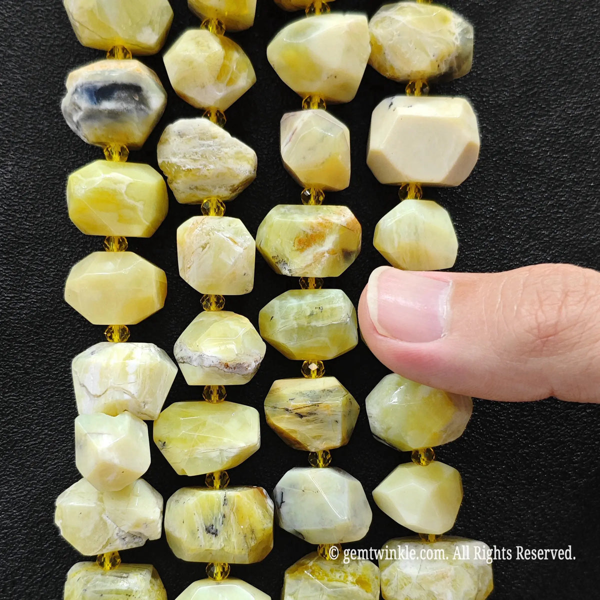 Faceted natural stone beads in pale yellow, cream, and white with subtle gray and brown veining, strung on gold cord against a black background, with a finger pointing at the beads.