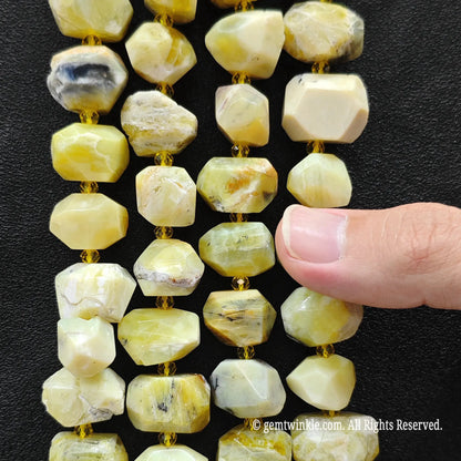 Faceted natural stone beads in pale yellow, cream, and white with subtle gray and brown veining, strung on gold cord against a black background, with a finger pointing at the beads.