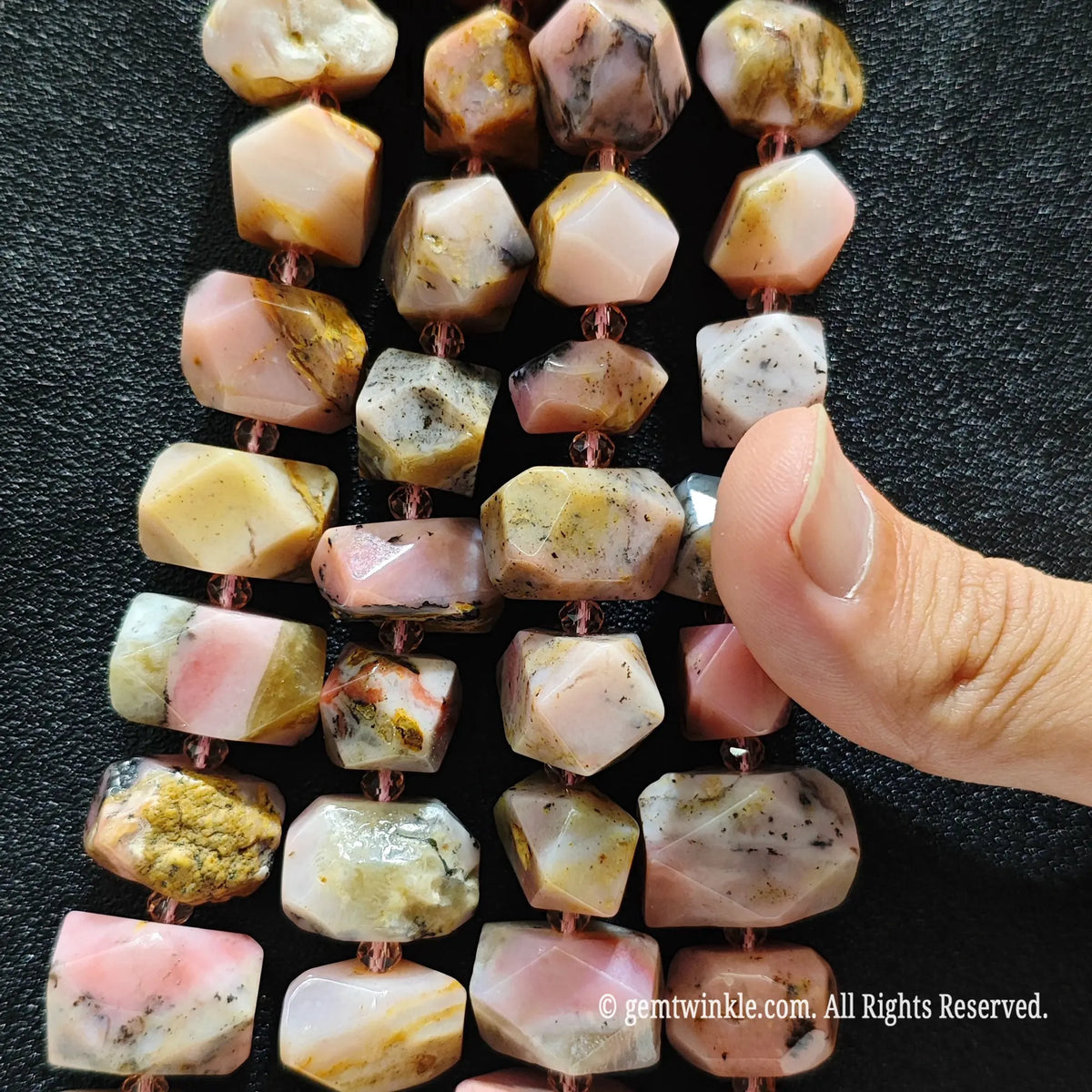 Multiple strands of faceted pink chrysocolla gemstone beads with copper-colored cord, displayed on a black textured background with a hand pointing to the beads.