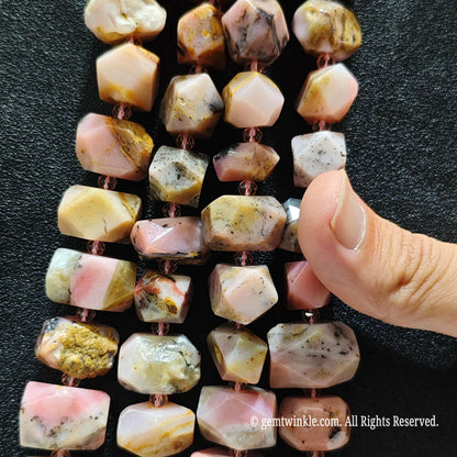 Multiple strands of faceted pink chrysocolla gemstone beads with copper-colored cord, displayed on a black textured background with a hand pointing to the beads.