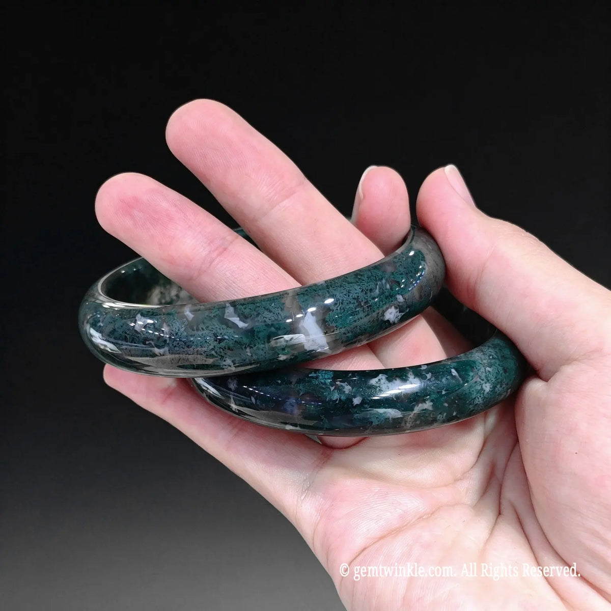 A pair of polished dark green gemstone bangles with white inclusions, held in a person's hand against a dark background.