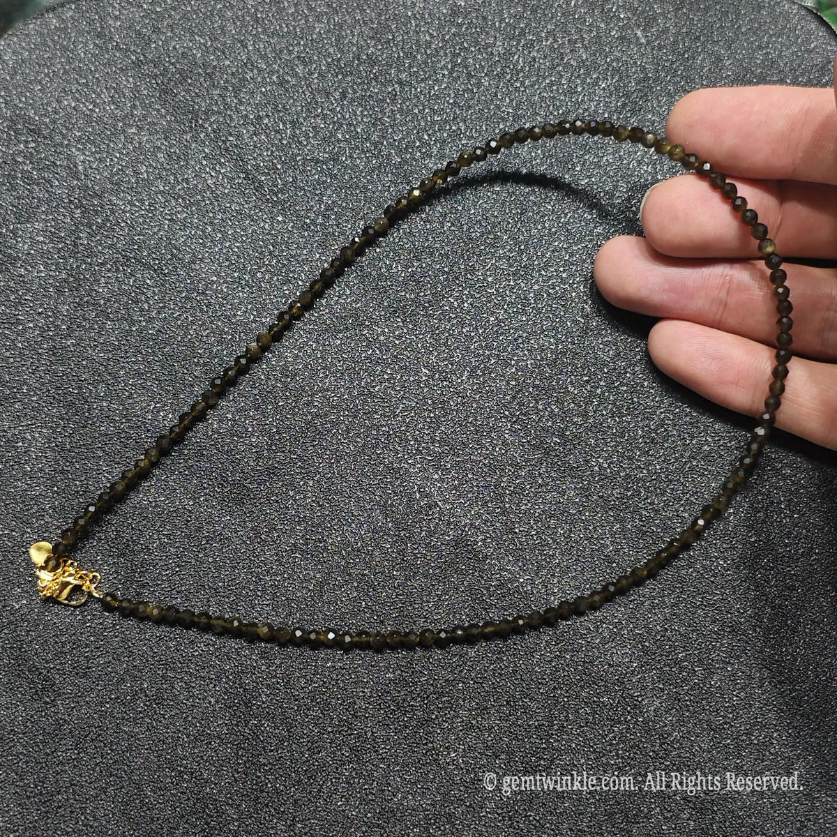 Hand holding 3mm micro faceted Gold Sheen Obsidian necklace strand showing deep black color and golden sheen