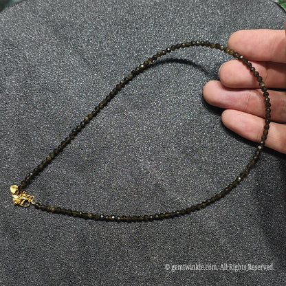 Hand holding 3mm micro faceted Gold Sheen Obsidian necklace strand showing deep black color and golden sheen