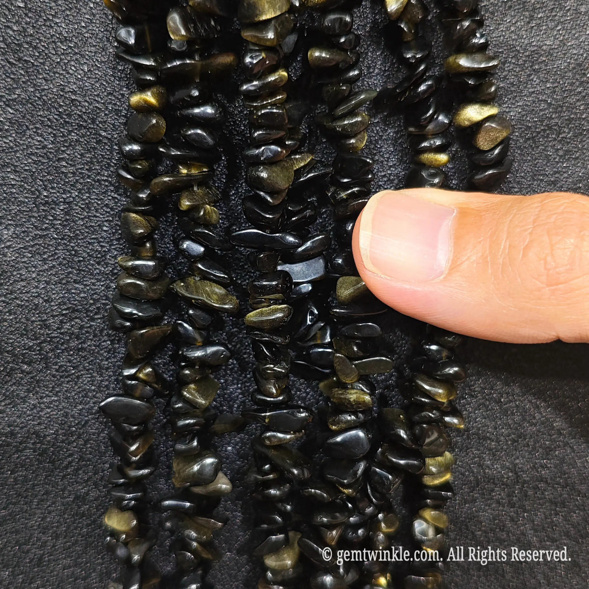 Fingers holding shimmering natural golden obsidian gemstone chip beads on black background[cite: 9]