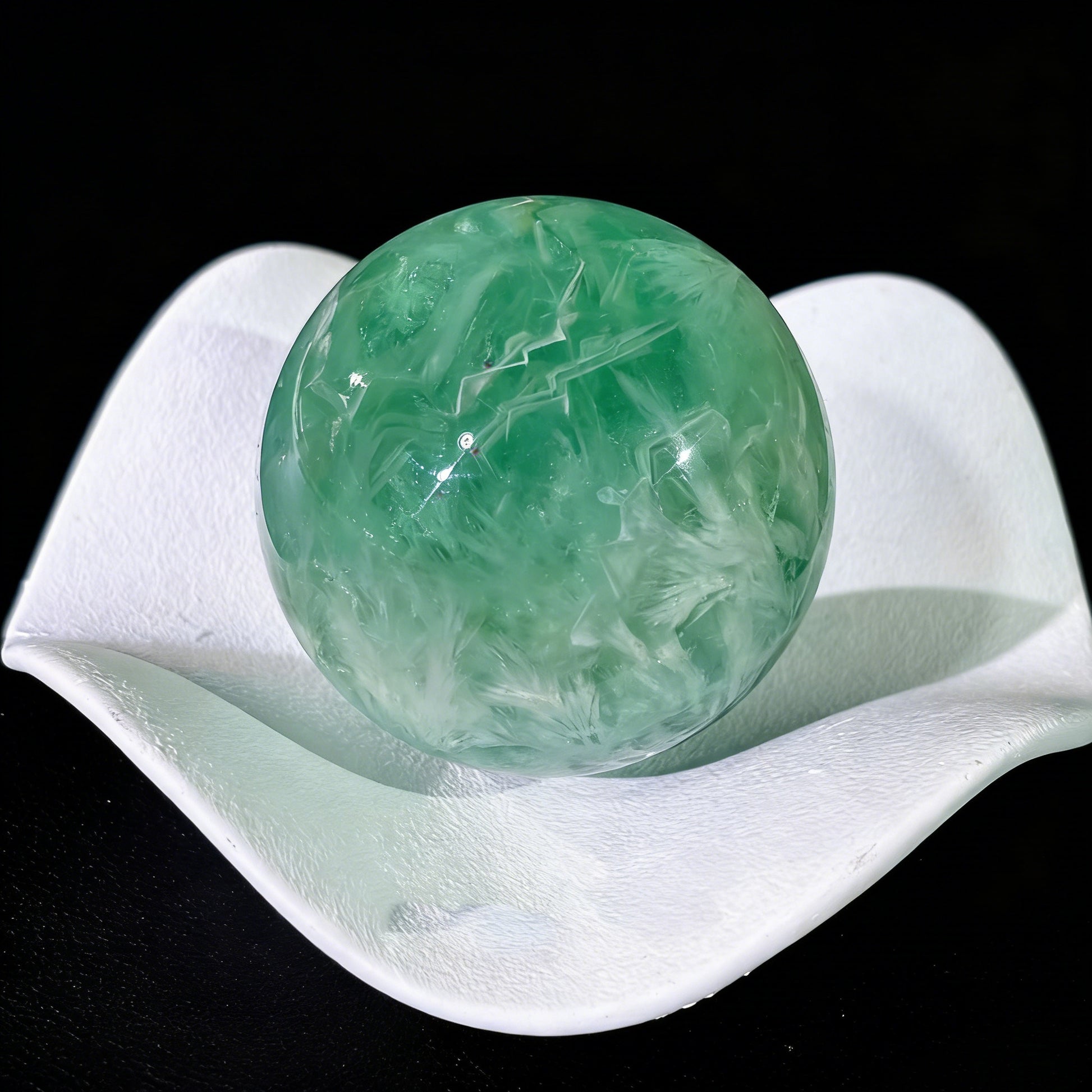 Green fluorite sphere with feather inclusions resting on white ceramic dish, showcasing translucency and internal patterns
