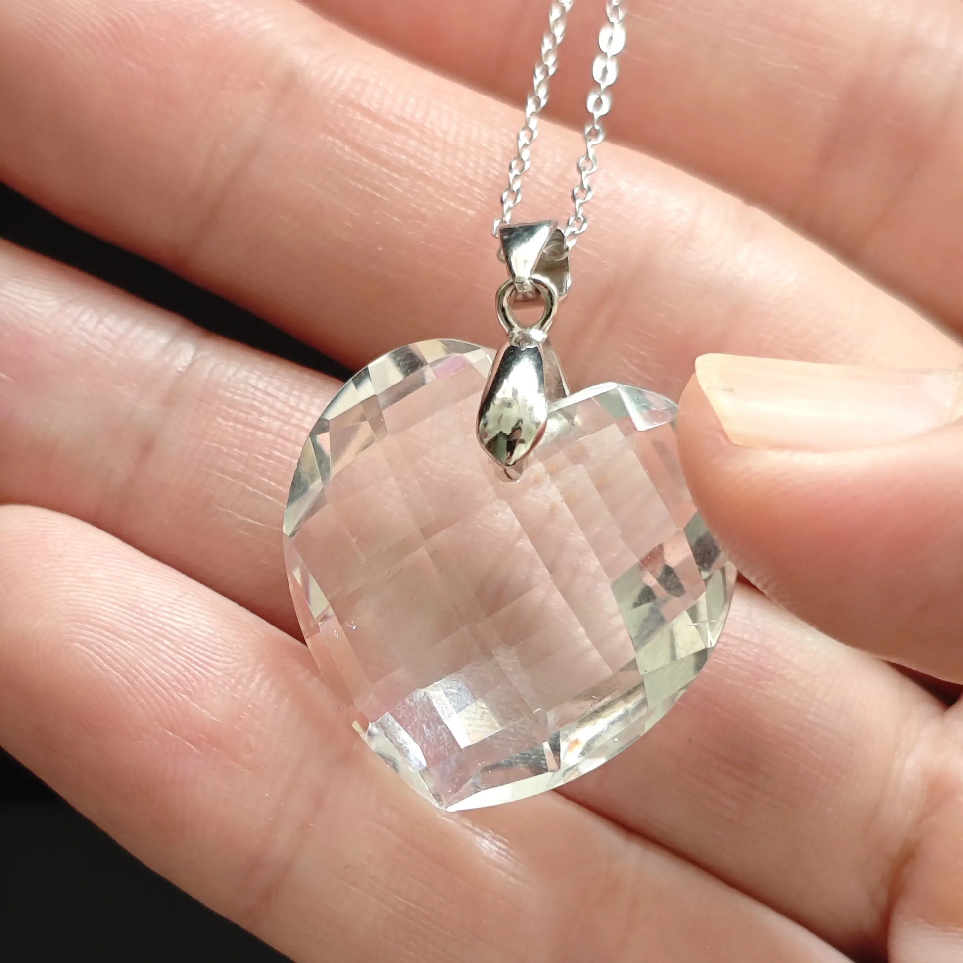 Hand holding clear quartz heart pendant, front-facing view highlighting multi-faceted cut and internal light play through transparent crystal
