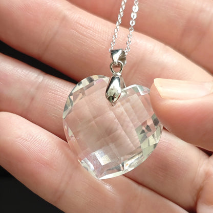 Hand holding clear quartz heart pendant, front-facing view highlighting multi-faceted cut and internal light play through transparent crystal