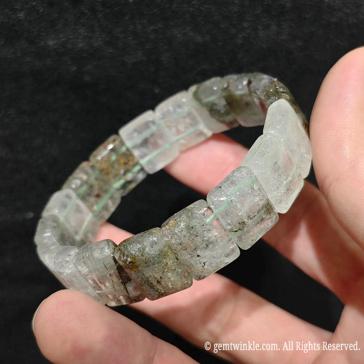 A beaded stretch bracelet made of chunky, translucent pale greenish-gray gemstone cubes, held in a person's hand against a dark textured background.