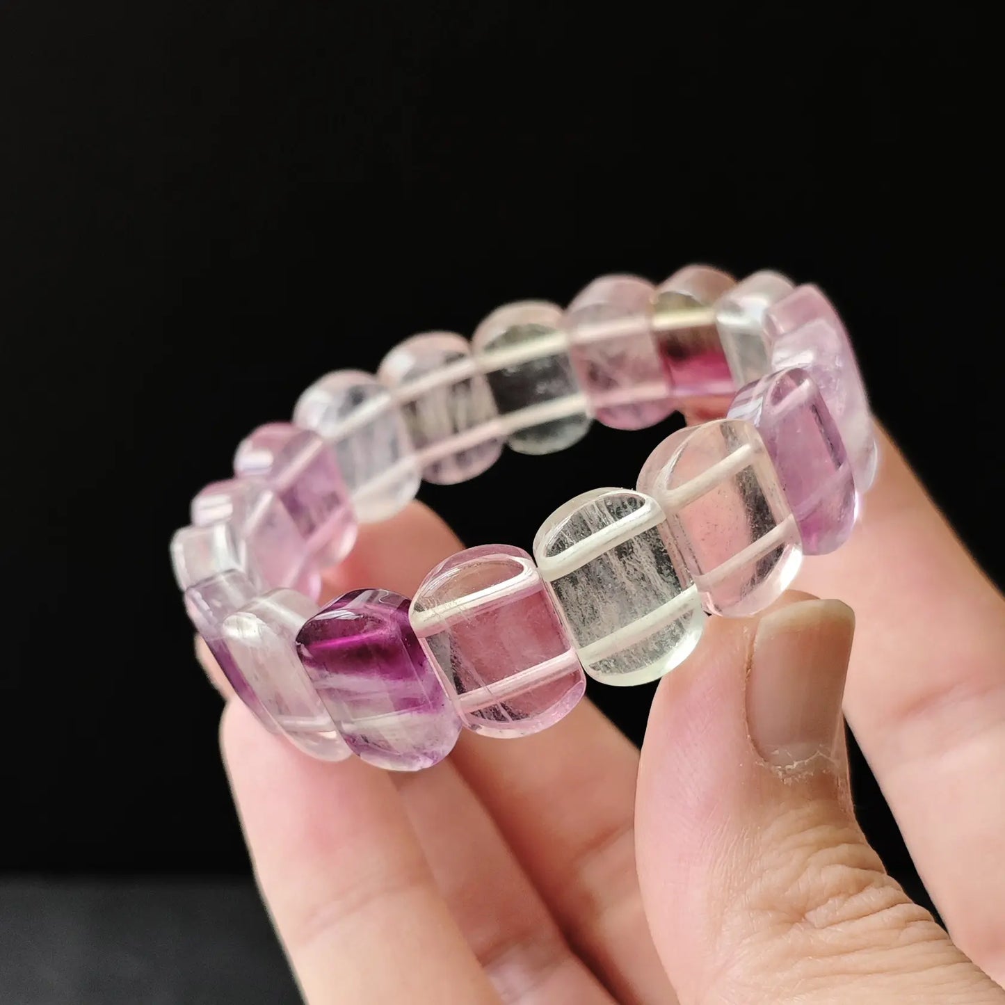 Natural multicolor fluorite oval bead bracelet held in hand, showing purple-to-pink-to-clear gradient and faceted surface on black background