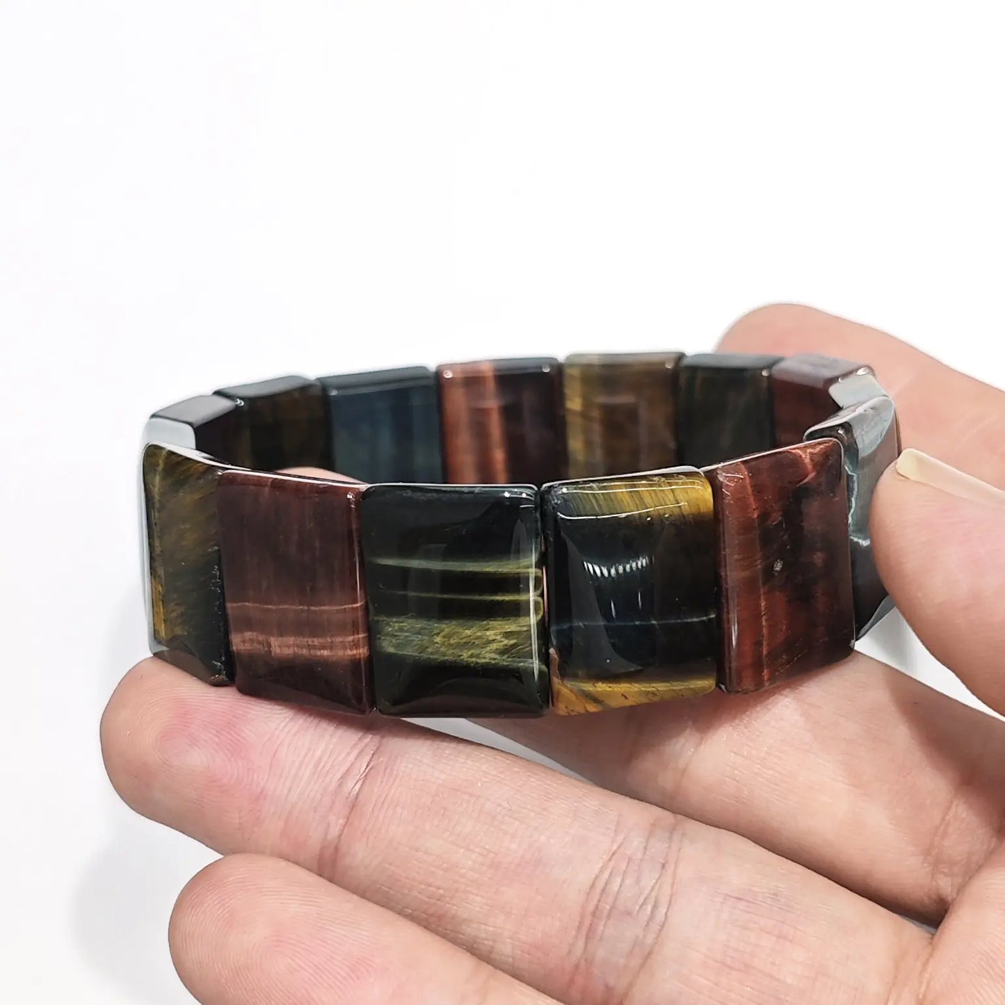 Hand holding natural multicolor tiger eye elastic bangle with rectangular beads (red, yellow, blue, black bands), showcasing chatoyant silk-like sheen on white background