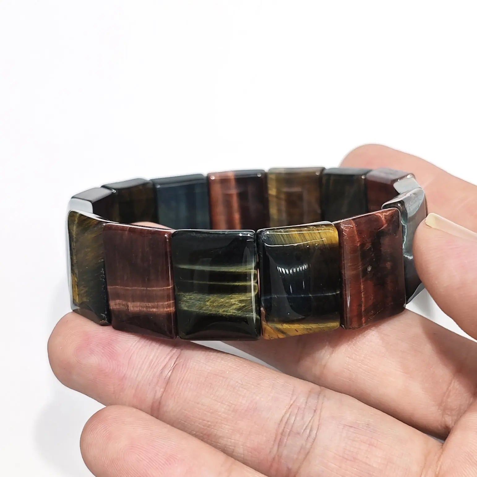 Hand holding natural multicolor tiger eye elastic bangle with rectangular beads (red, yellow, blue, black bands), showcasing chatoyant silk-like sheen on white background