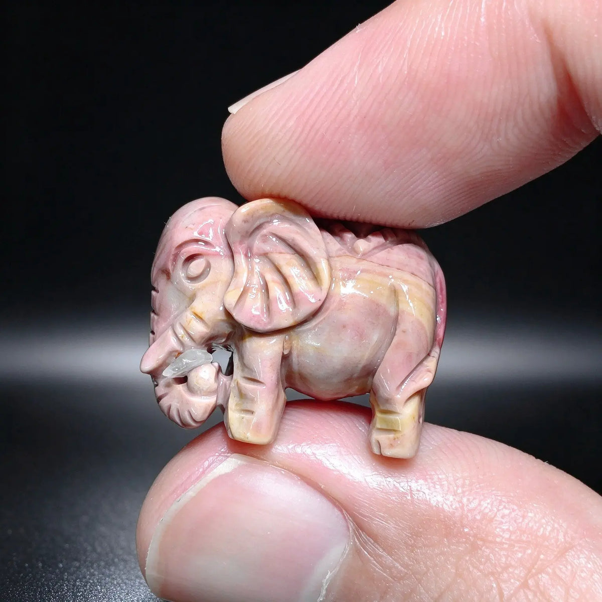 Finger holding a dusty rose natural Alxa agate mini elephant carved statue with curled trunk showing smooth polish