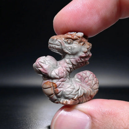 Finger holding a tiny natural Alxa agate dragon holding ingot carved mini statue showcasing pink gray natural color zoning
