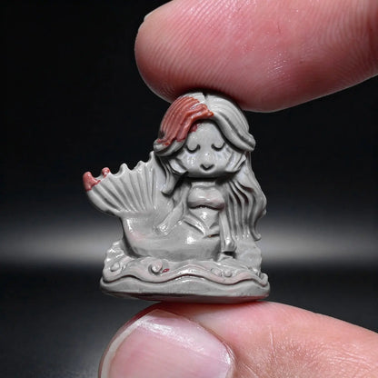Finger holding a tiny natural Alxa agate mermaid carved mini statue with gray body and reddish hair highlights
