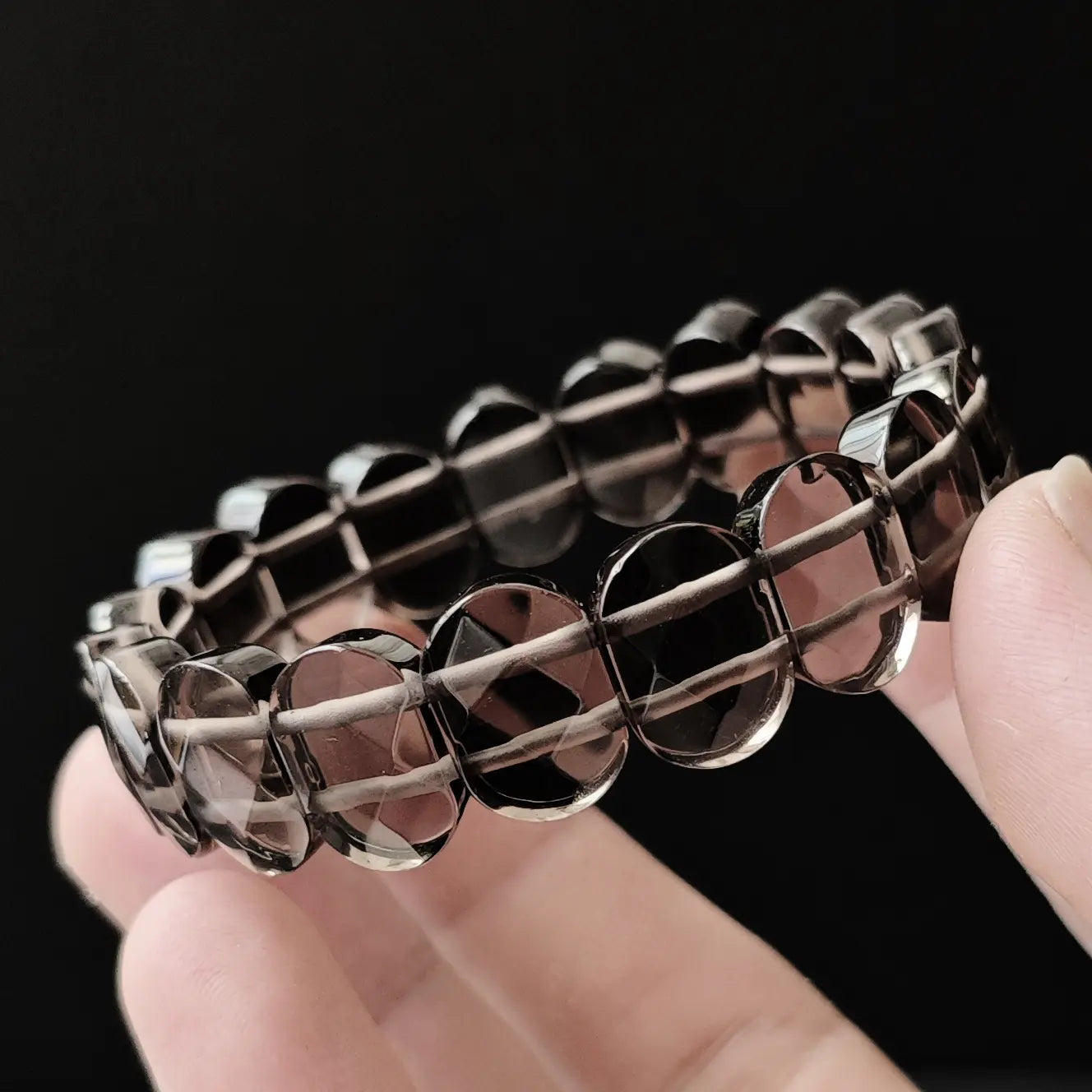 Natural ice-grade black obsidian faceted bracelet with oval beads, high transparency and glassy luster, held in hand on black background; premium spiritual jewelry for women, energy protection accessory