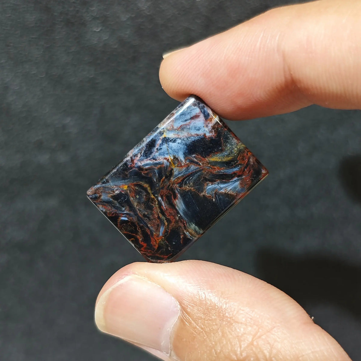 Large rectangular natural petersite loose stone block held in fingers showing blue and gold storm chatoyant patterns