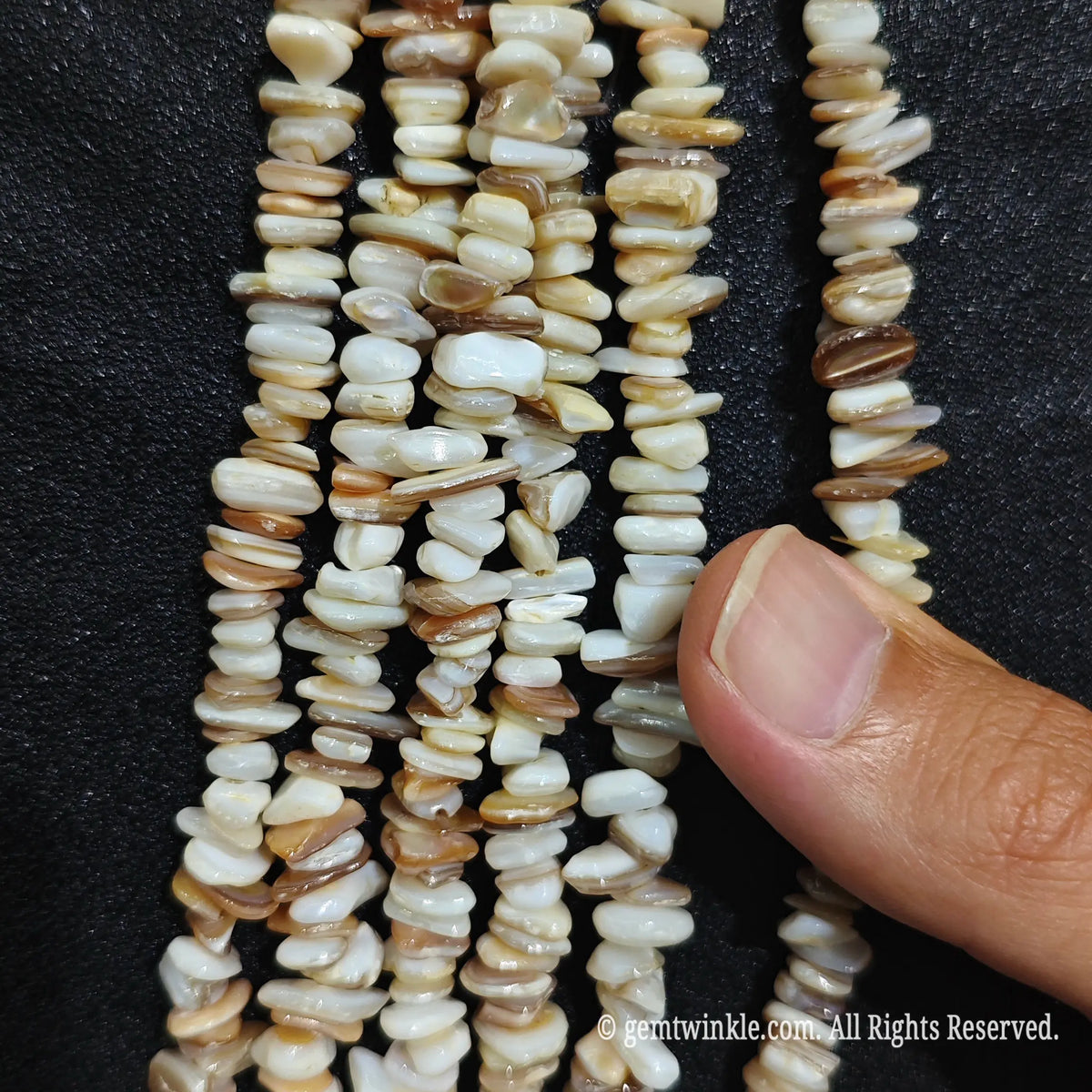 Fingers holding shimmering natural mother of pearl shell gemstone chip beads on black background[cite: 8]