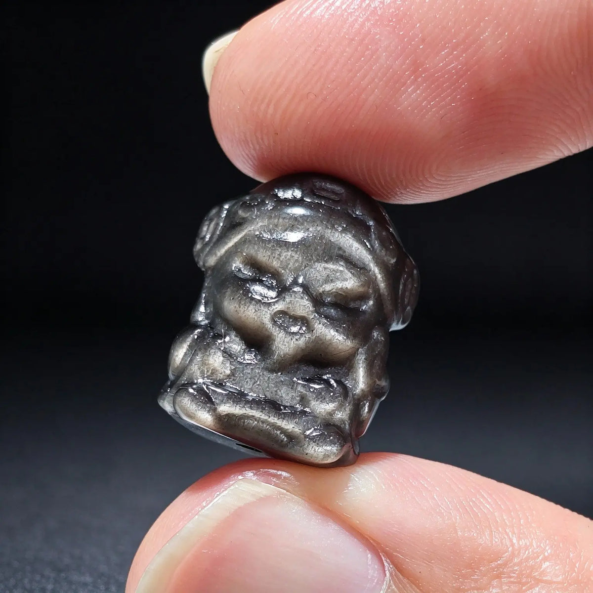 Fingers holding a small natural silver sheen obsidian God of Wealth carved focal bead showing metallic sheen reflection