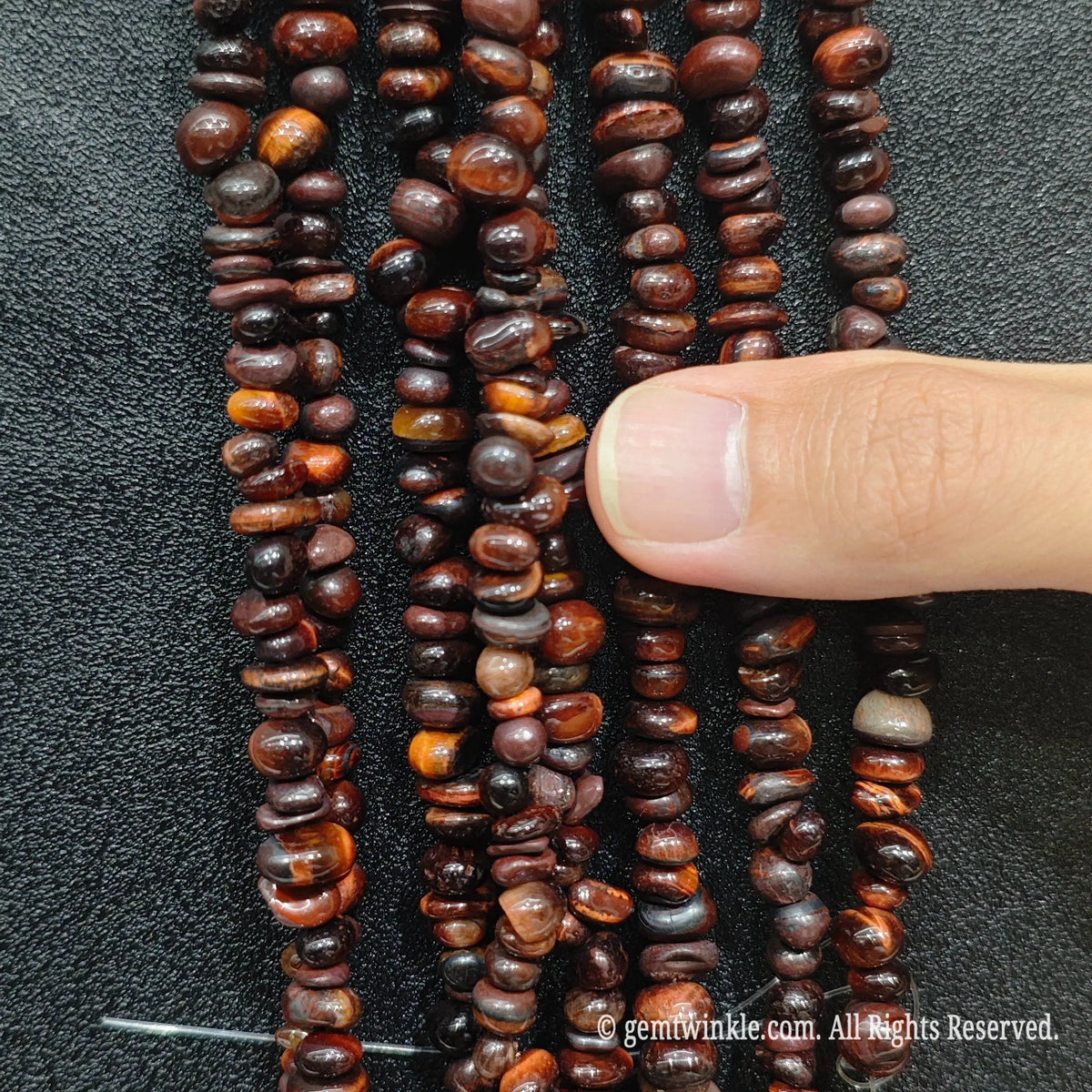 Fingers holding shimmering red tiger eye gemstone chip beads on black background