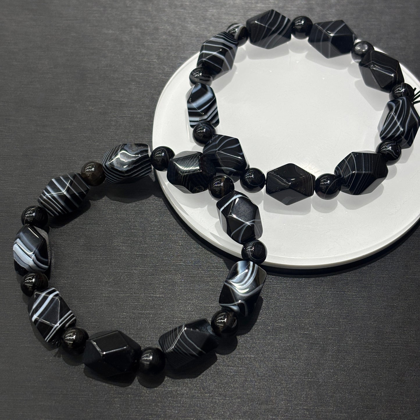 Two black and white banded agate irregular bead bracelets layered on white ceramic disc, minimalist geometric stacking