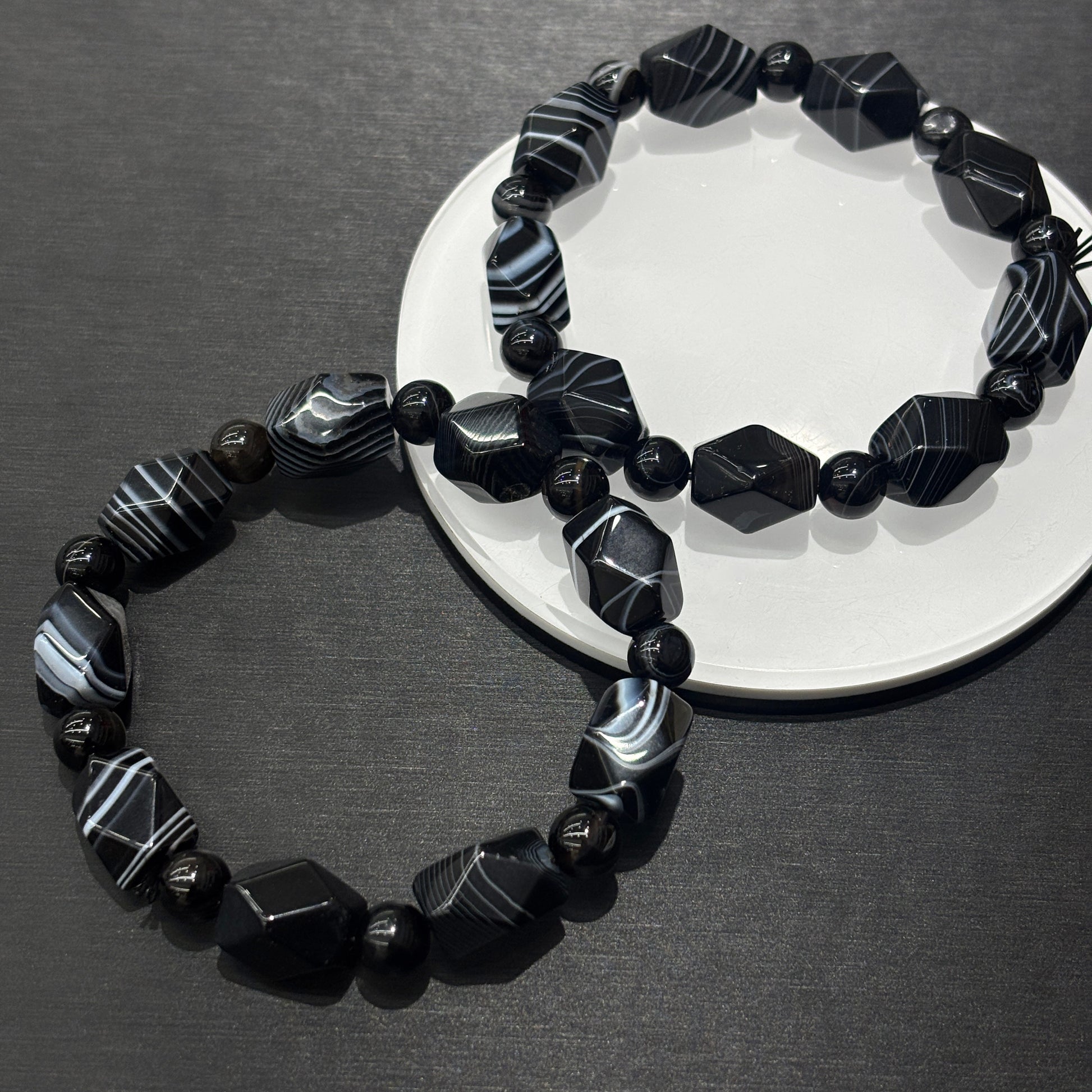 Two black and white banded agate irregular bead bracelets layered on white ceramic disc, minimalist geometric stacking