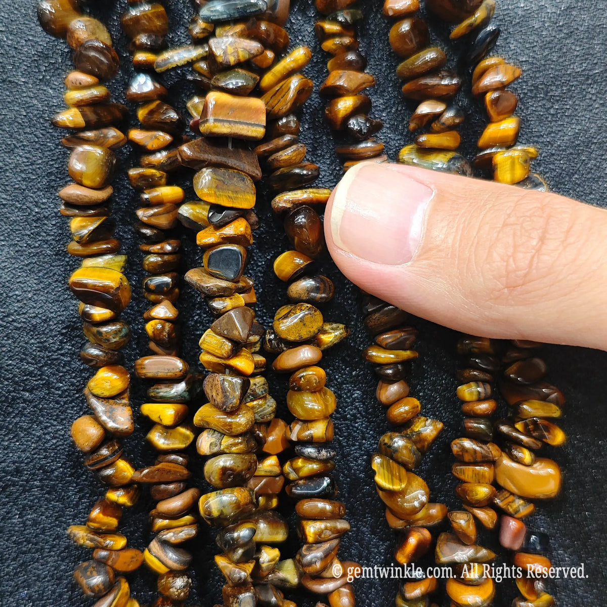 Fingers holding shimmering yellow tiger eye gemstone chip beads on black background[cite: 8]
