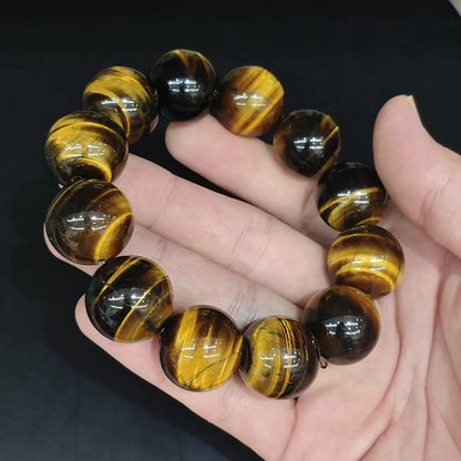 Hand holding a classic yellow tiger’s eye bracelet with uniformly polished spherical beads. Visible size range from small (≈6mm) to large (≈20mm), featuring deep brown base and vivid golden-yellow bands that catch the light.