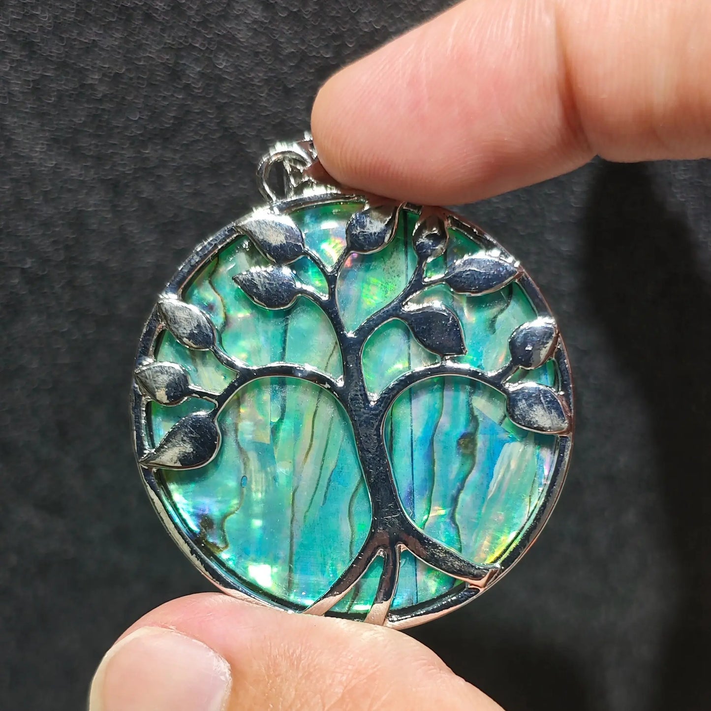 Side angle display of Tree of Life abalone shell pendant held in fingers showcasing smooth alloy rim finish