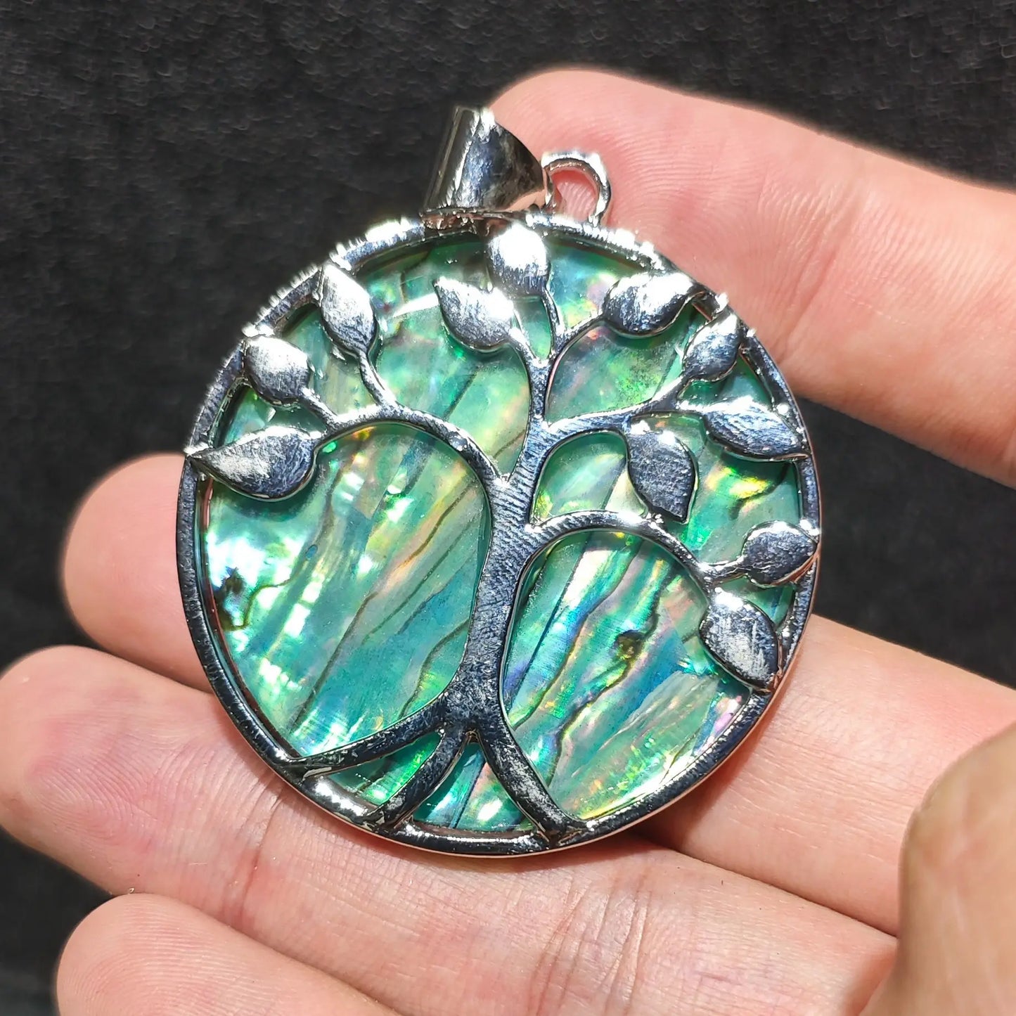 Close up view of 50mm Tree of Life abalone shell pendant held on palm showcasing green and peacock blue metallic luster