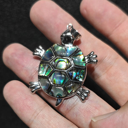 Angled close up view of sea turtle pendant displaying peacock blue and green abalone shell reflections held in hand