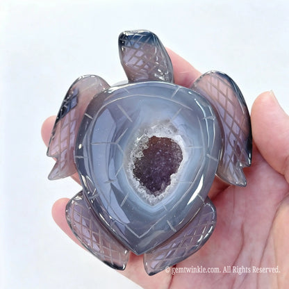 Single agate geode turtle figurine held in hand showing deep grey chalcedony shell and raw crystal cavity