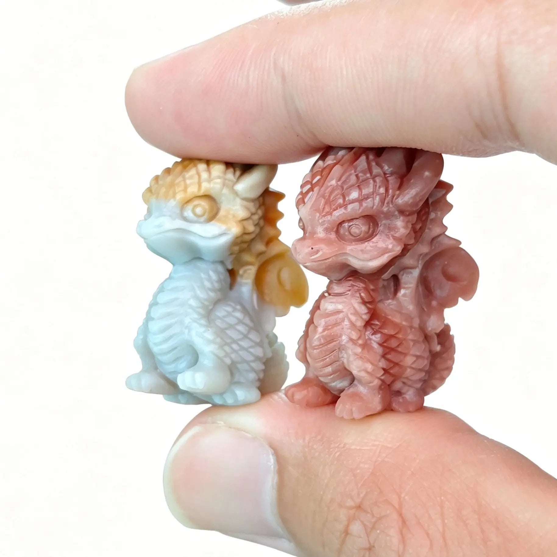 Main view of two mini Alashan agate carved winged dragon figurines held between fingers showing blue-yellow and terracotta red colors