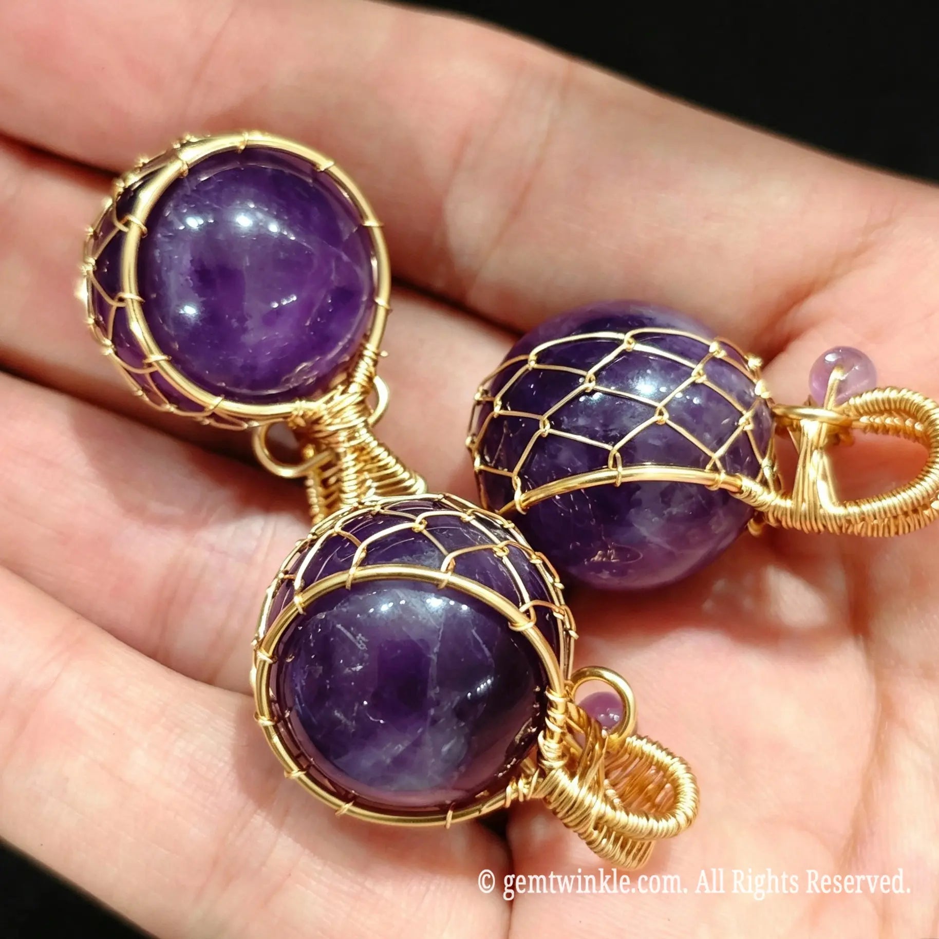 Three gold wire wrapped amethyst sphere pendants resting on palm showing rich purple color and wire mesh detail