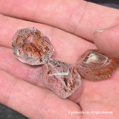 Handheld view of three black gold super seven asymmetrical faceted beads showing dense rutile and mica inclusion patterns