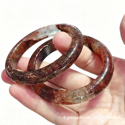 Pair of smooth round black gold super seven quartz bangles held in hand displaying rich rutile needle texture